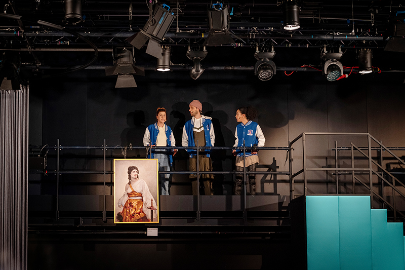 Sami (Estelle Schmidlin), Elvis (Frederic Lilje) und Bulli (Lola Merz Robinson)(v.l.n.r.) stehen auf der der Galerie der JES-Bühne. Die drei Schauspieler*innen unterhalten sich. Am Geländer hängt das Gemälde "Judith" von August Riedel. Rechts ist die Showtreppe zu sehen, die nach unten auf die Bühne führt. Links sieht man den Rand des Fadenvorhangs der die Band umgibt.