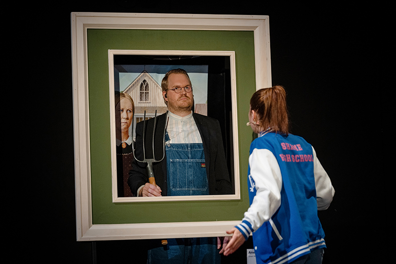 Sami (Estelle Schmidlin) steht vor dem Gemälde "American Gothic" von Grant Wood. Der Mann im Gemälde wird dargestellt vom Schlagzeuger Philipp Bindreiter, der mit einer Mistgabel in der Hand in einem grünen Bilderrahmen steht. Sami spricht zum Gemälde. Auf dem Rücken ihrer blauen College-Jacke steht der Schriftzug "SHAME HIGH SCHOOL"