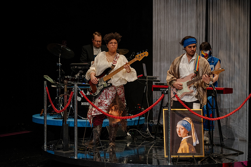 Die Band, bestehend aus Fermin Emanuel Abanto Ulloa (Gitarre), Philipp Bindreiter (Schlagzeug), Clara John (Klavier) und Lonteshia Jayne Stripf (Bass), steht auf einem silbernen Podest. Alle sind als Gemälde/Kunstwerke dargestellt. Im Hintergrund ist ein weißer Fadenvorhang.