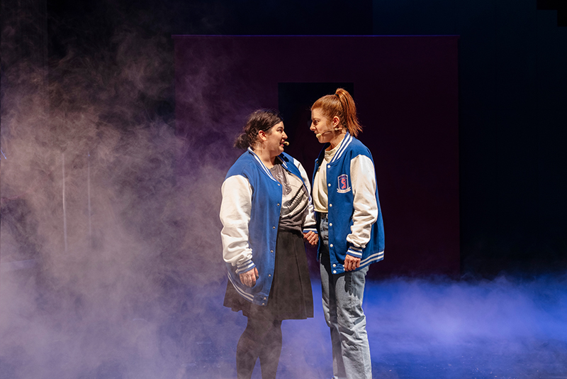 Sami (Estelle Schmidlin) und Birte (Daniela Ruocco) stehen sich gegenüber, schauen sich verliebt an und halten sich an der Hand. Beide haben blaue College-Jacken an und sind von Nebel umgeben. 